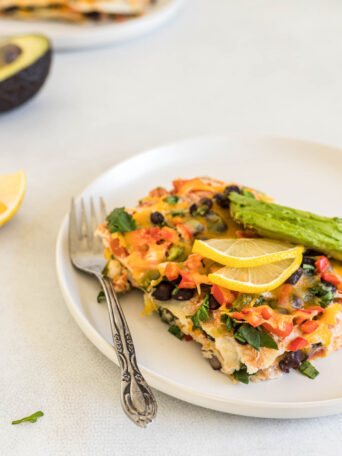 A big slice of Vegetarian Mexican Lasagna is on a plate with lemon slices and avocado on top. A fork sits next to it. Some lemon and avocado, as well as another plate of lasagna sit in the background.