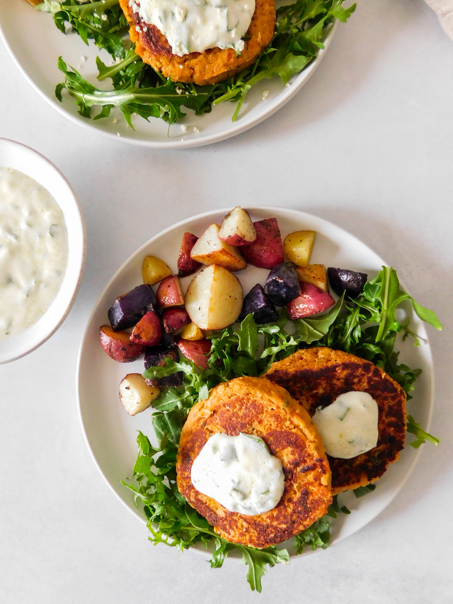 An overhead view of Sun-Dried Tomato Chickpea Patties on two plates with arugula and roasted potatoes.