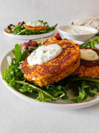 A Sun-Dried Tomato Chickpea Patty sits on top of another. They both are topped with a yogurt sauce and are sitting on top of a bed of arugula. Another plate with patties sits in the background, along with a small bowl with yogurt sauce.