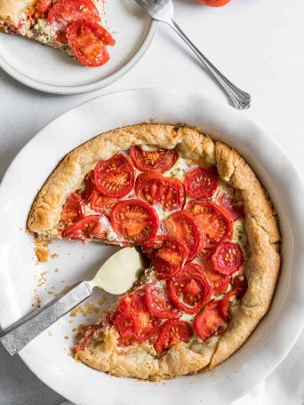 An overhead view of a tomato tart with a slice cut out of it. The slice sits on a small plate to the side with a fork.