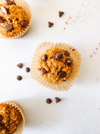 A Healthy Oat Flour Pumpkin Muffin with chocolate chips on top of it sits on top of a muffin liner that is frayed out. Two other muffins sit near by and chocolate chips are scattered around.