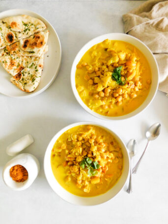 Two bowls of Cauliflower Potato Curry With Chickpeas. Naan bread sits to the side along with two spoons, a linen cloth, and a mortar and pestle with curry powder in it.