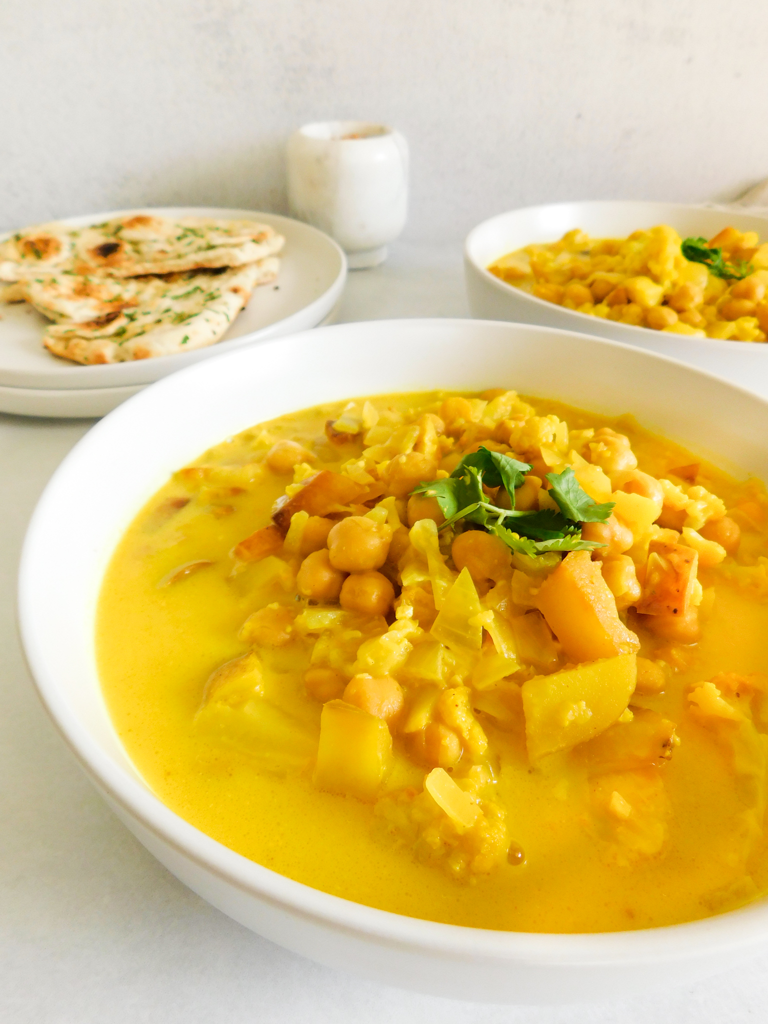 A side angle of Cauliflower Potato Curry with naan bread, another soup bowl, and a pestle in the background.