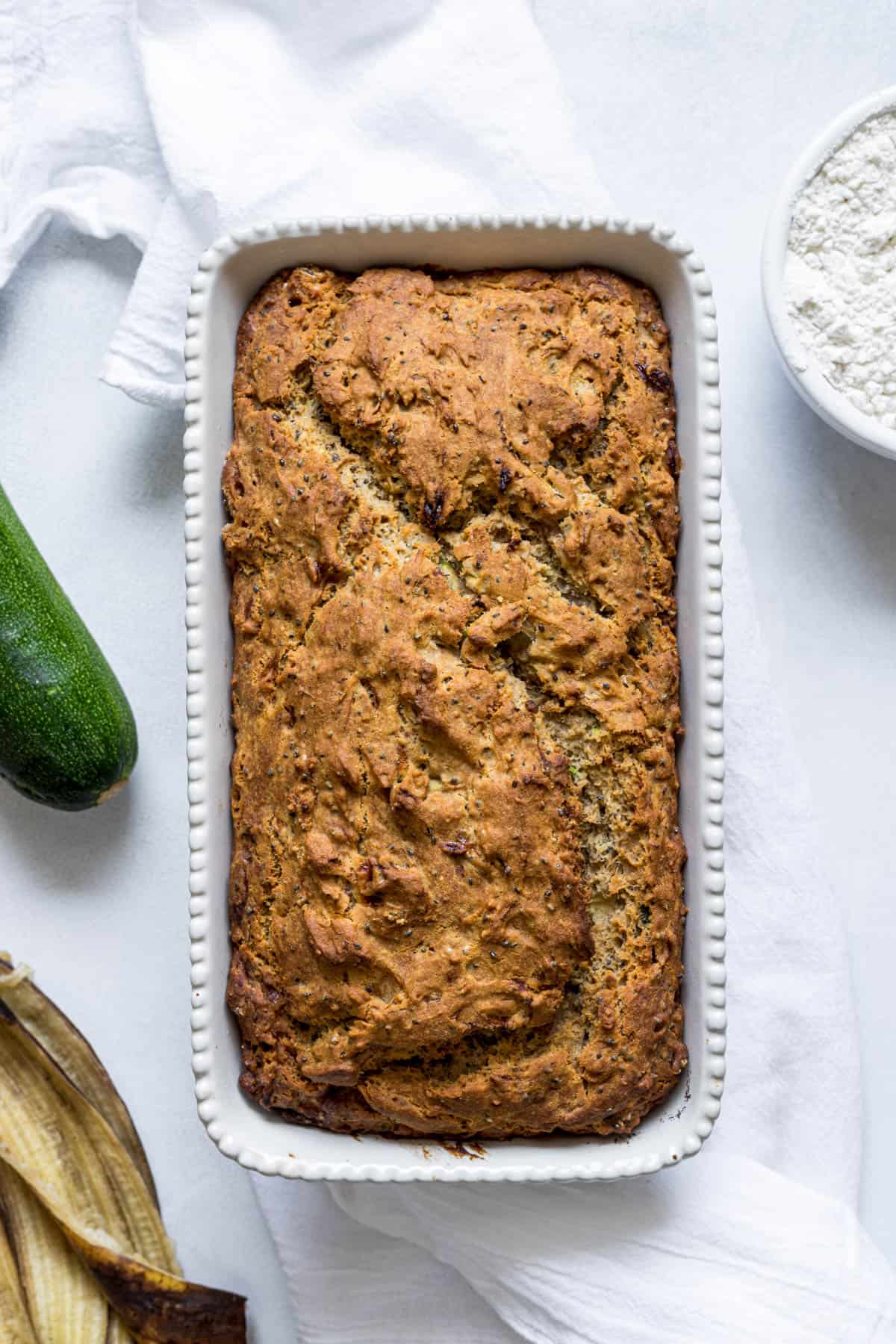 A loaf of zucchini banana bread on a dish towel. Zucchini, a banana peel, and flour sit next to the loaf.