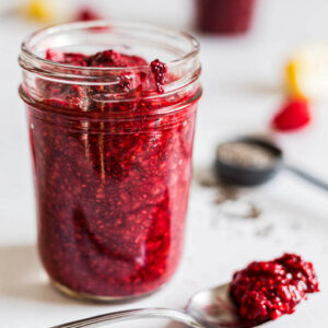 Side view of raspberry chia jam in a jar. A spoon with jam on it lies beside it.