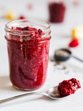 Side view of raspberry chia jam in a jar. A spoon with jam on it lies beside it.