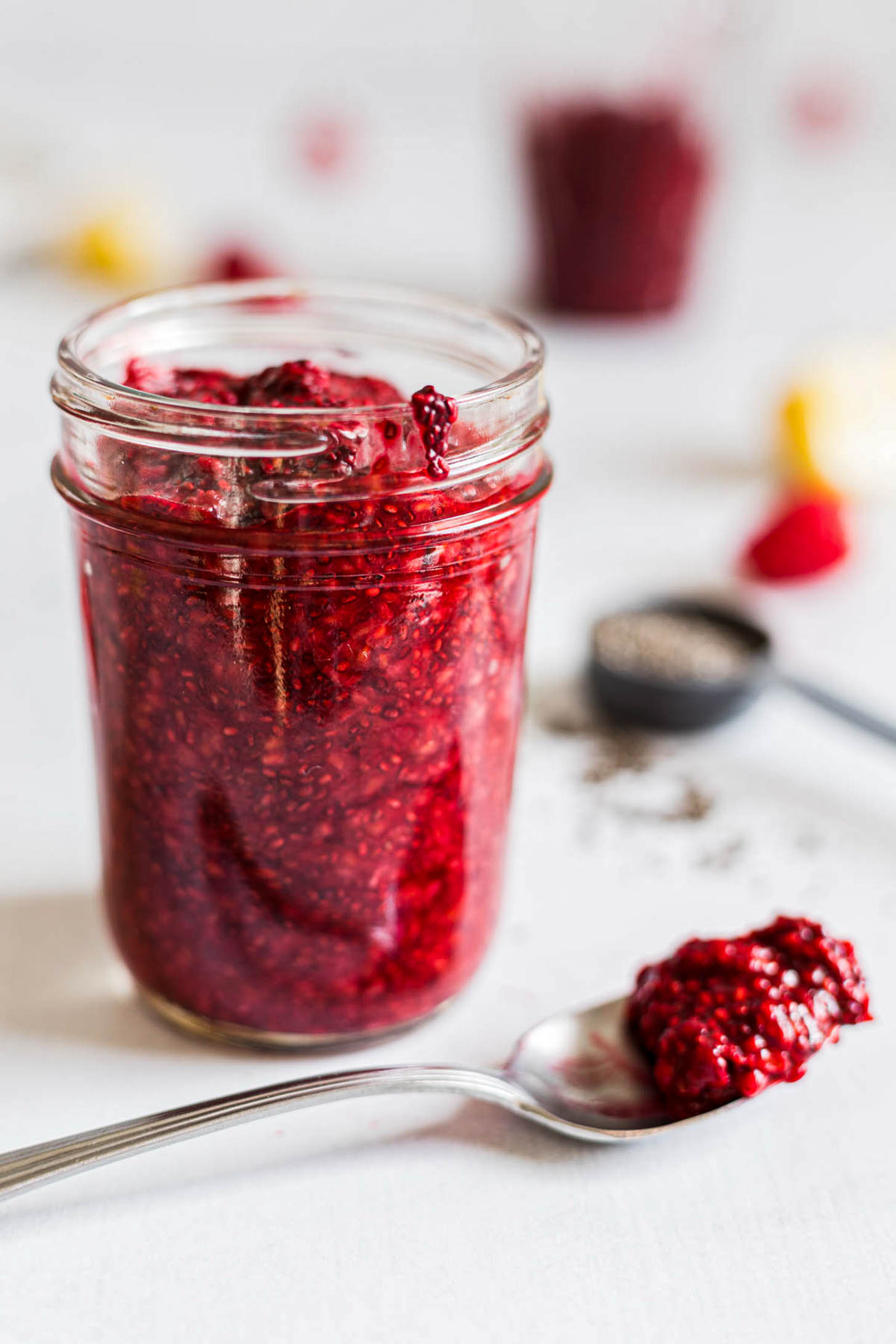 Side view of raspberry chia jam in a jar. A spoon with jam on it lies beside it.