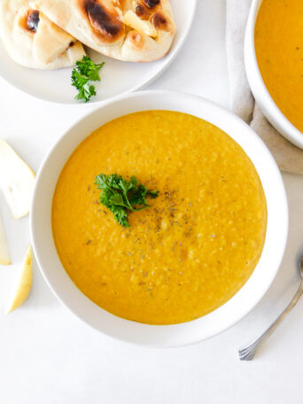 An overhead shot of a bowl of Curried Butternut Squash and Pear Soup with sprig of parsley on it. Naan bread and pear slices surround the soup.