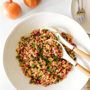 A large serving bowl filled with Fall Lentil Farro Salad With Pomegranate. Two serving spoons sit in the bowl. Two pomegranates sit off to the side along with a plate and two forks.