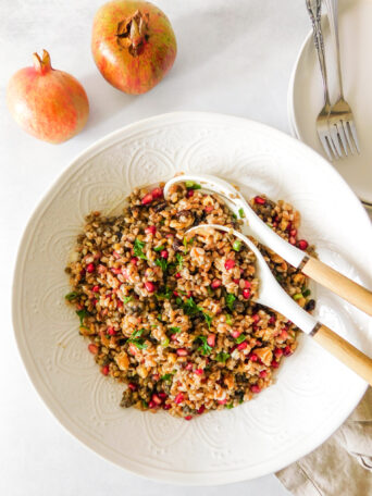 A large serving bowl filled with Fall Lentil Farro Salad With Pomegranate. Two serving spoons sit in the bowl. Two pomegranates sit off to the side along with a plate and two forks.