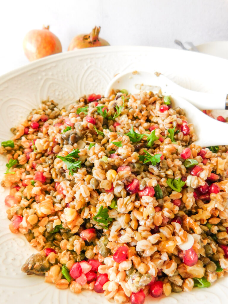 A bowl of farro lentil salad with two serving spoons in the bowl. Two pomegranates are sitting behind the bowl.