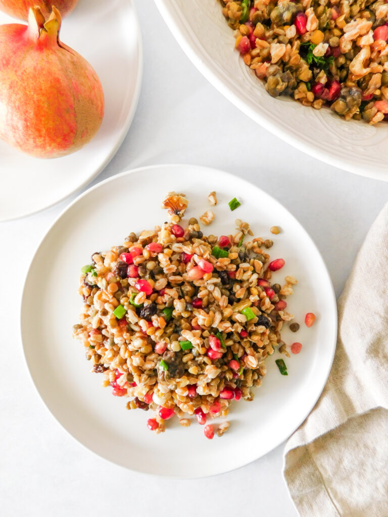 A small plate with Fall Lentil Farro Salad With Pomegranate. A plate with a pomegranate sits off to the side as well as a large serving bowl with the remainder of the salad. A dishcloth sits next to the main plate.