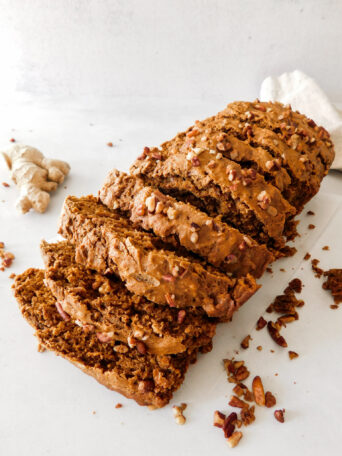 A Vegan Gingerbread Loaf is sitting sideways that has been cut into slices. A piece of ginger sits in the background.