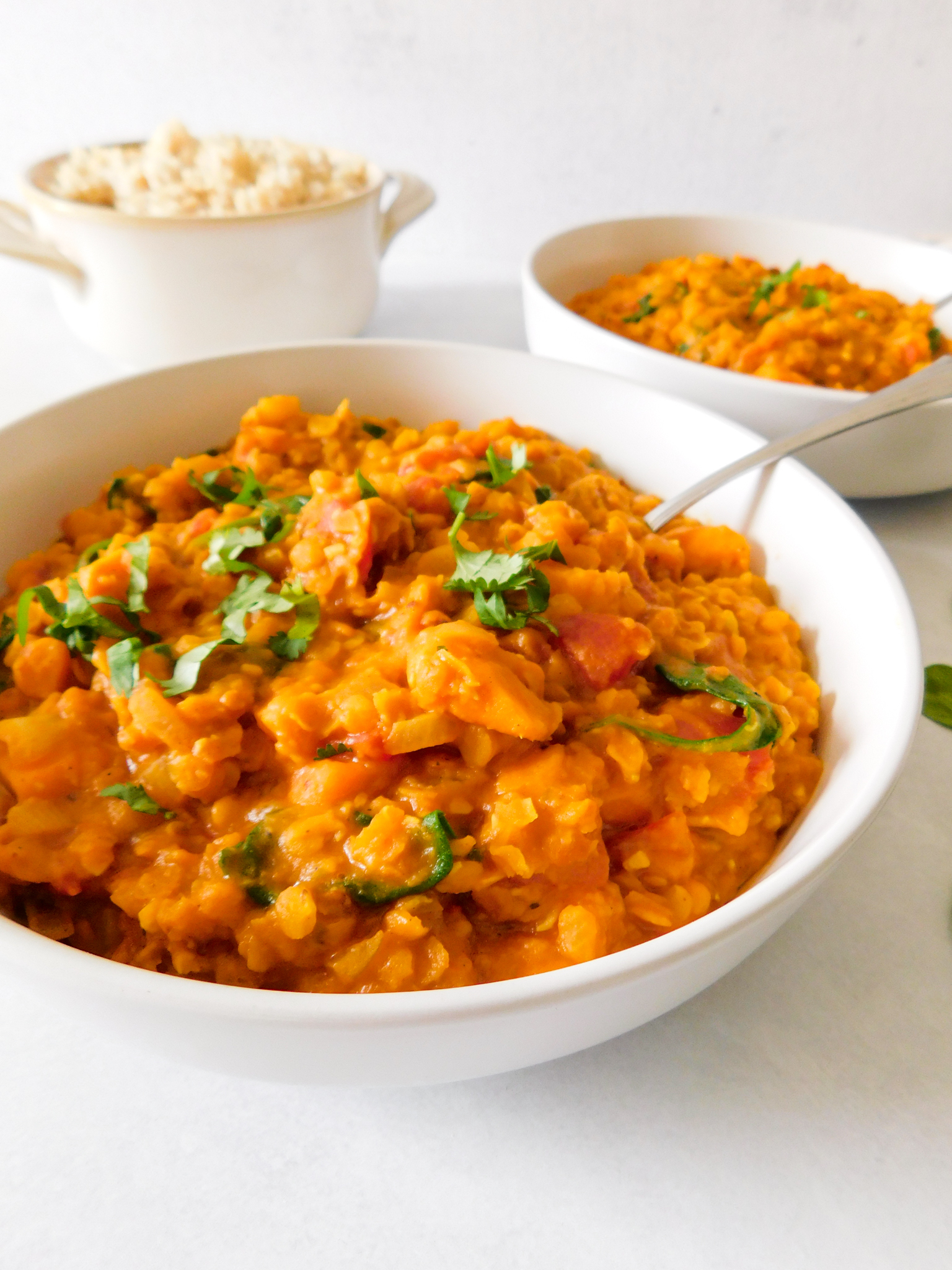 A side view of red lentil curry with another bowl of curry in the background as well as a bowl of rice.