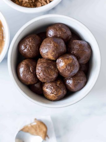 A bowl of Chocolate Almond Balls with a spoon of peanut butter next to it and a bowl of cocoa powder.