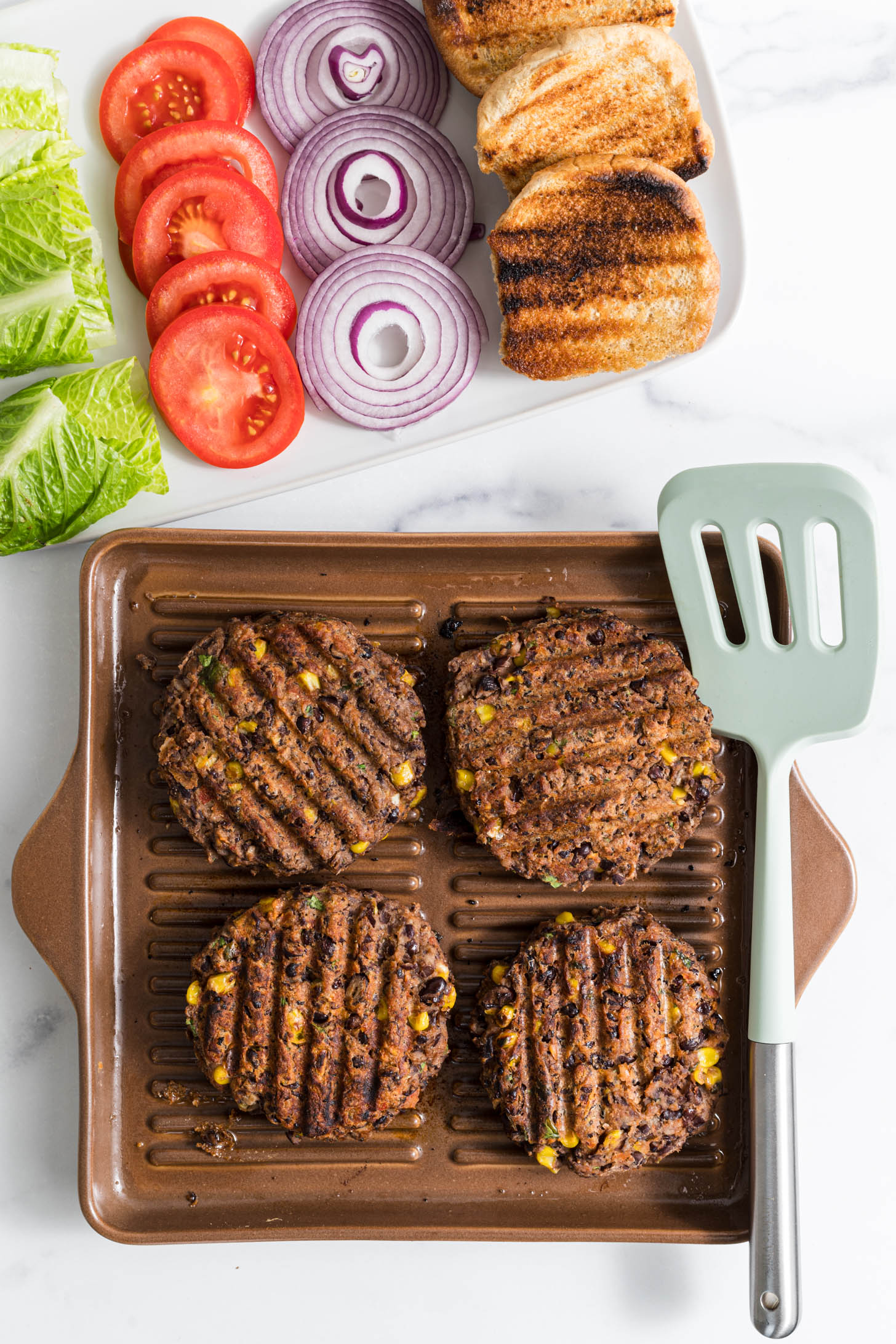 Cooked burgers on grill pan with burger toppings laid out next to it.