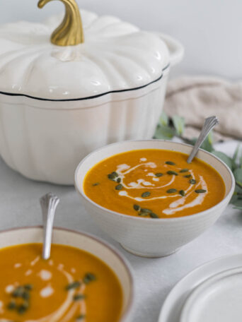 Two bowls of Carrot Ginger Sweet Potato Soup with a Pumpkin Dutch Oven behind them.